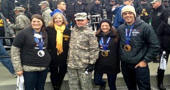 A group of people, including individuals in military uniforms and others wearing medals, posing for a photo in an outdoor setting with a crowd in the background.