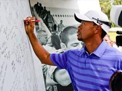 A man in a purple striped shirt and white cap writes on a large whiteboard with a red marker, standing in front of a black-and-white photograph.